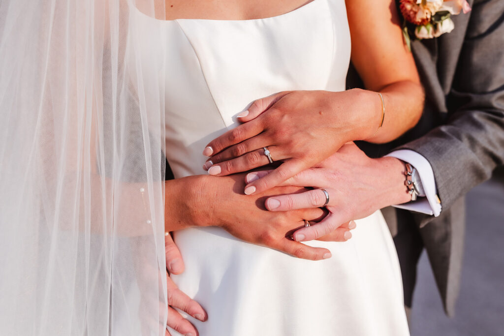 A close-up of a bride and groom embracing, showing their hands with wedding rings resting on the bride’s white dress. The bride wears a veil and holds a bouquet, while the groom wears a suit.