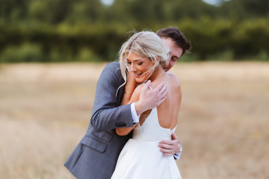 A bride in a white dress smiles and leans into the embrace of a groom in a gray suit, standing together in a sunlit outdoor field with trees in the blurred background.