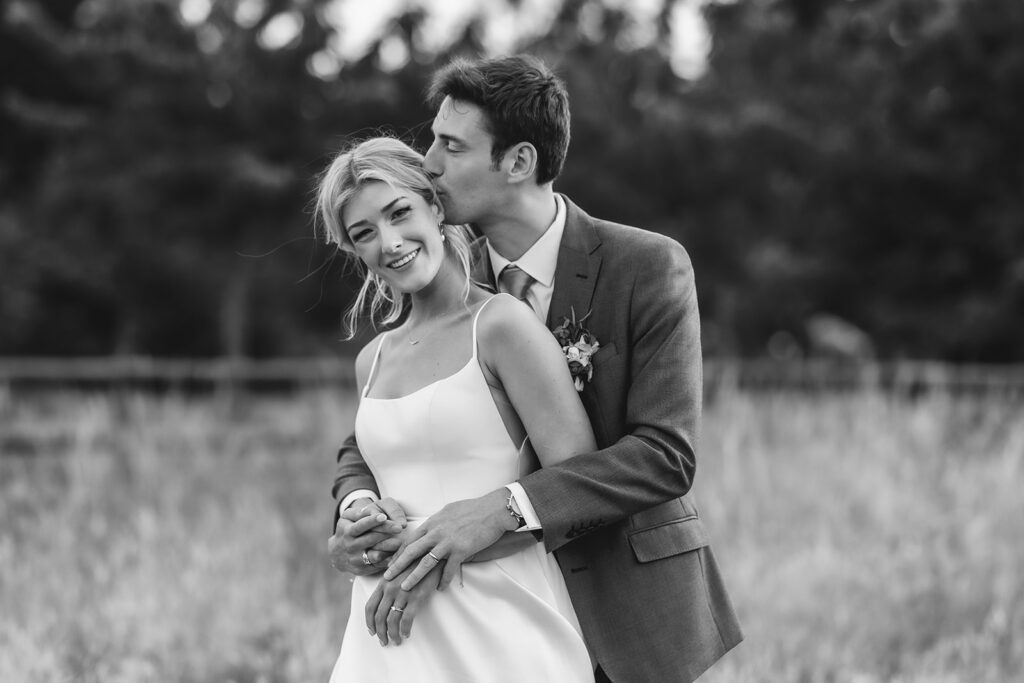 A groom in a suit embraces and kisses the forehead of a smiling bride in a white dress outdoors, with tall grass and trees in the background. The photo is in black and white.