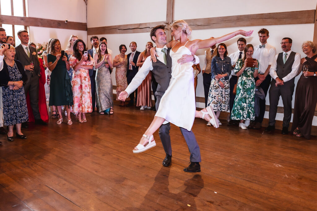 A couple dances joyfully in the center of a wooden-floored room, surrounded by smiling guests dressed in formal and semi-formal attire, watching and celebrating the moment.