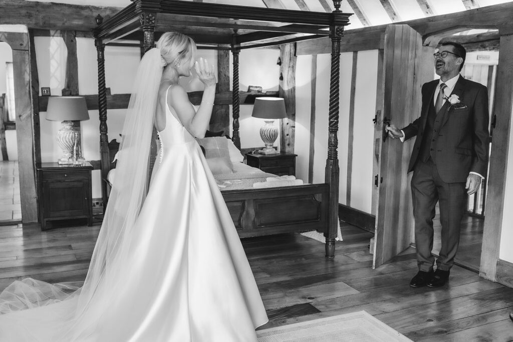 A bride in a white gown and veil happily greets an older man in a suit, likely her father, who stands in a doorway smiling. They are in a rustic, warmly lit bedroom with wooden beams and a four-poster bed.