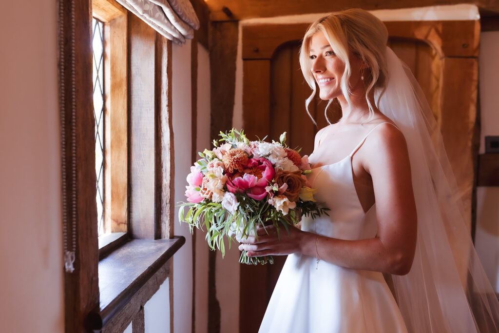 A bride in a white dress and veil holds a colorful bouquet of flowers while smiling and looking out of a sunlit window in a rustic wooden room.