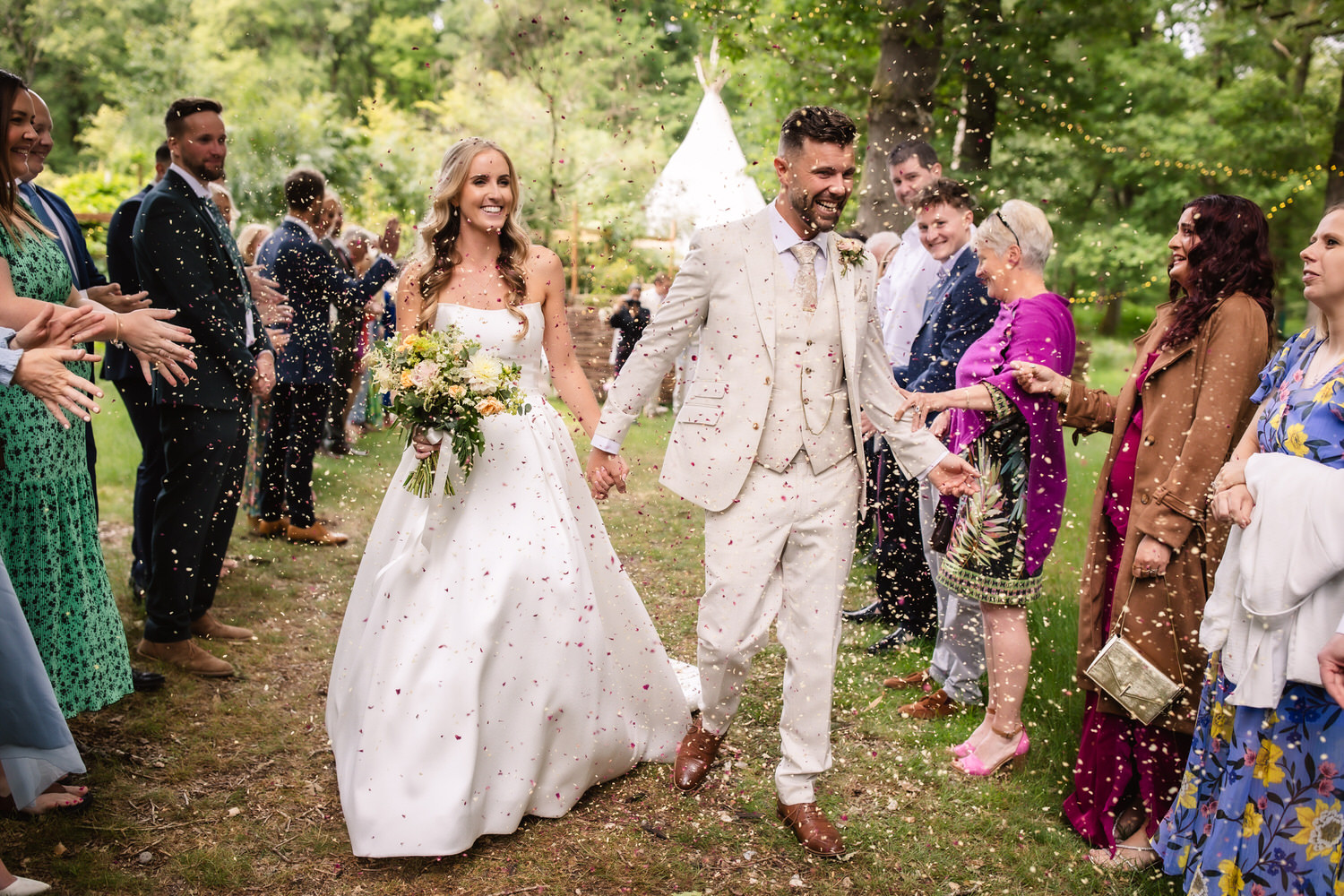 A bride and groom walk hand in hand outdoors, smiling as guests shower them with confetti. Captured by a Hampshire wedding photographer, the couple is surrounded by friends and family celebrating on a grassy path beside a white tent.