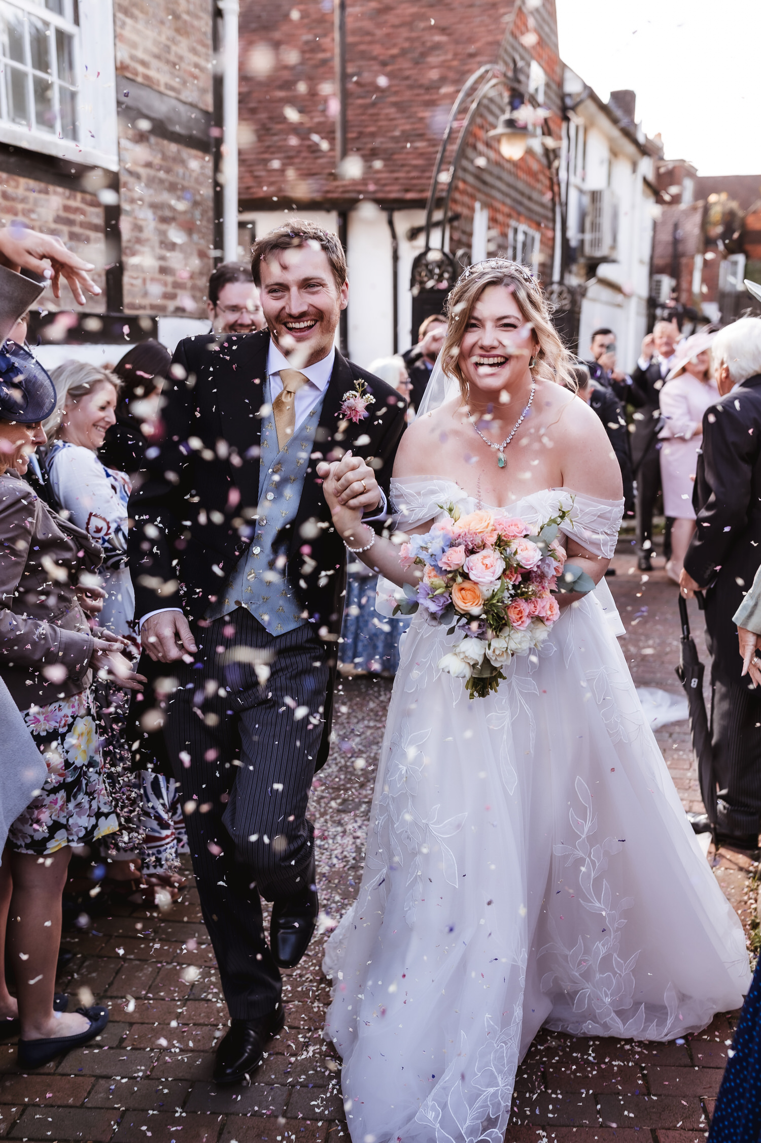 A newlywed couple, smiling and holding hands, walks through a crowd of guests throwing confetti. The bride wears a white dress and holds a colorful bouquet; the groom is in a suit with a light blue vest.