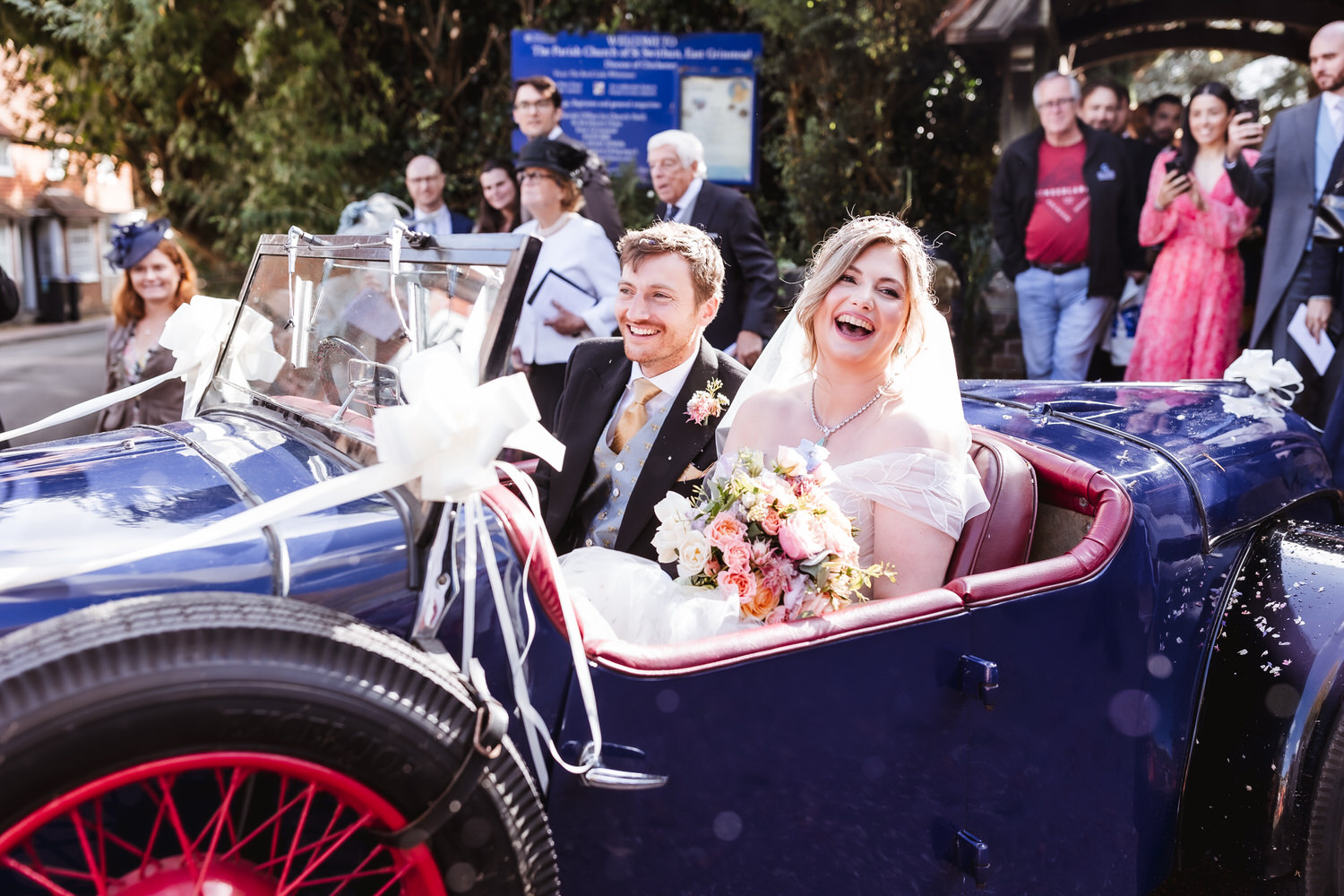 A joyful bride and groom sit in a vintage blue car decorated with white ribbons. The bride holds a bouquet and smiles widely, while guests stand cheering in the background on a sunny day.