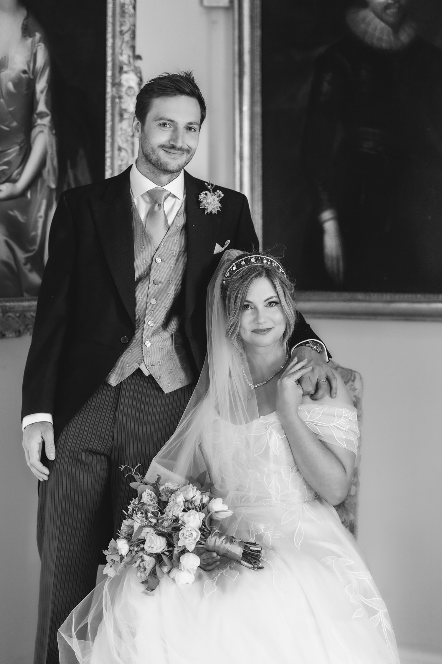 A smiling groom in a suit stands beside a seated bride in a wedding dress holding a bouquet, his hand on her shoulder. The photo is black and white, and they are posed in front of classical portraits.