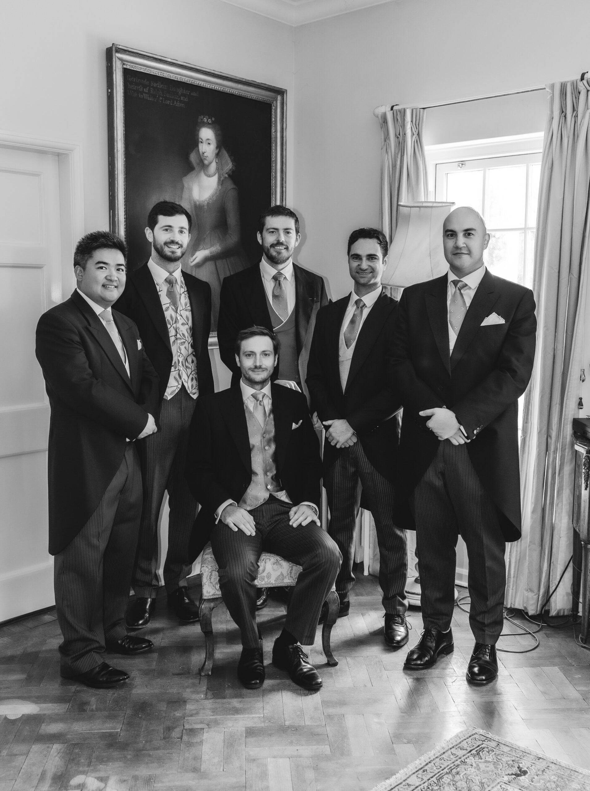Six men in formal suits pose for a group photo in a room with a large portrait on the wall and a window with curtains. One man is seated while the others stand around him, all smiling. The photo is in black and white.