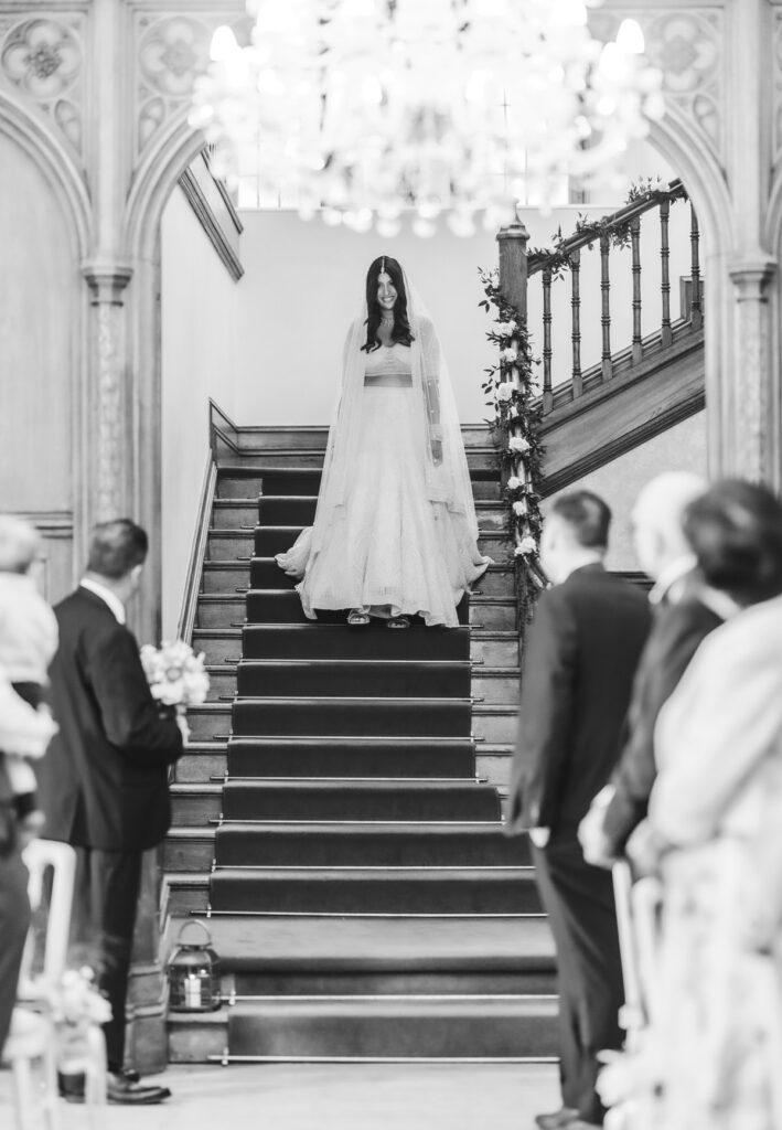 A bride in a long, flowing dress stands at the top of a staircase, looking down. Guests stand on either side of the aisle, watching her. The scene is set in an ornate, elegant venue. The image is black and white.