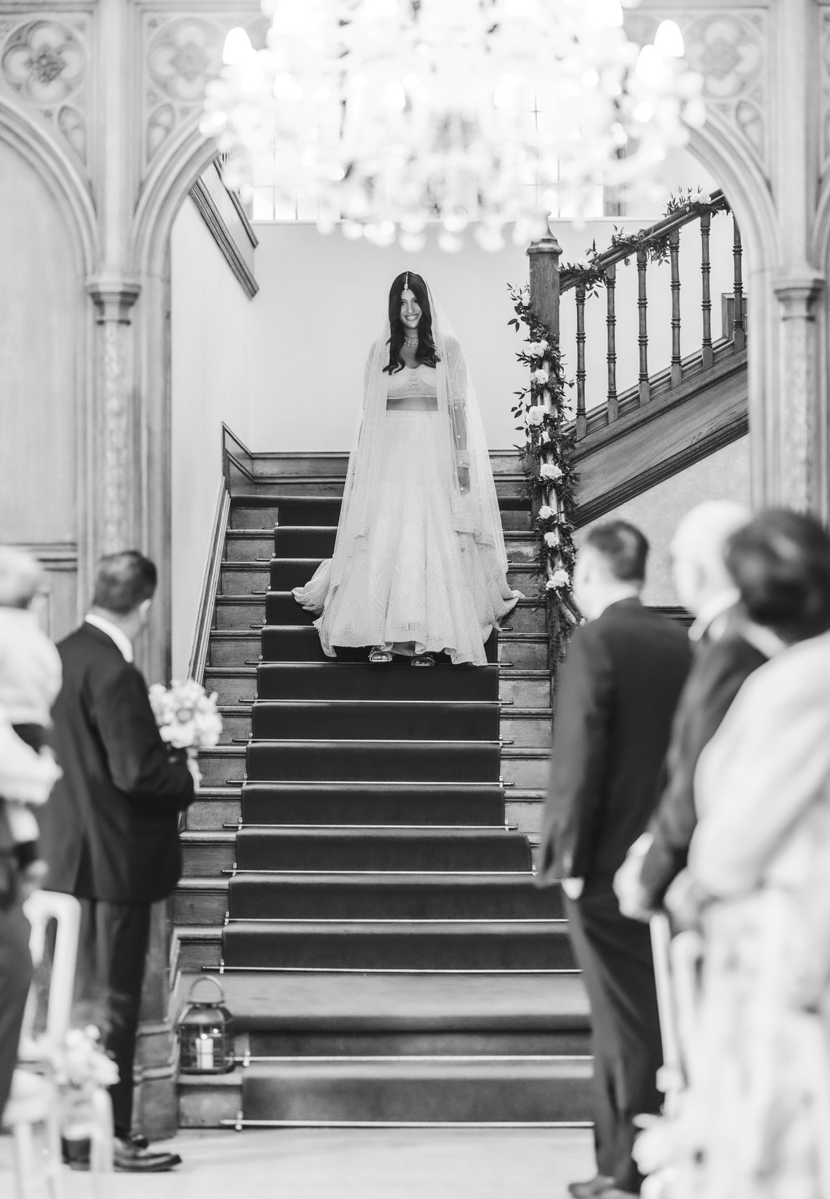 A bride in a long, flowing dress stands at the top of a staircase, looking down. Guests stand on either side of the aisle, watching her. The scene is set in an ornate, elegant venue. The image is black and white.