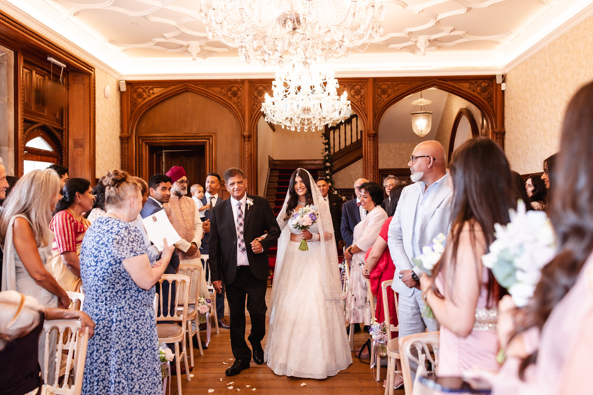 A bride in a white gown walks down the aisle holding a bouquet, escorted by an older man, as guests seated on either side watch and smile in a beautifully decorated indoor venue with a chandelier.
