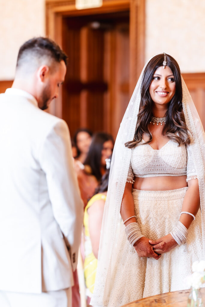 A bride in a white traditional outfit and veil smiles at a groom in a light suit during a wedding ceremony, with guests visible in the softly lit background.
