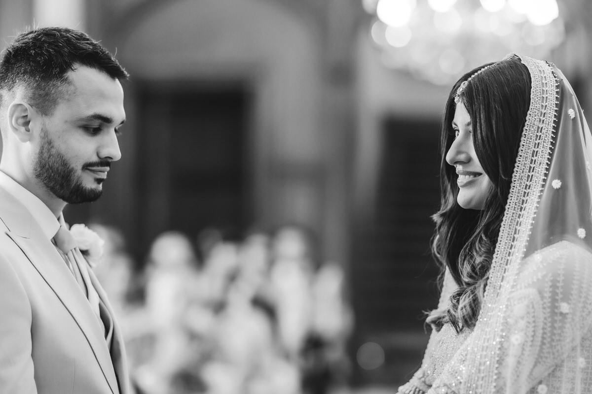 A bride and groom face each other during a wedding ceremony. The groom wears a light-colored suit, while the bride smiles, dressed in a traditional outfit with a decorative veil. The background is softly blurred.