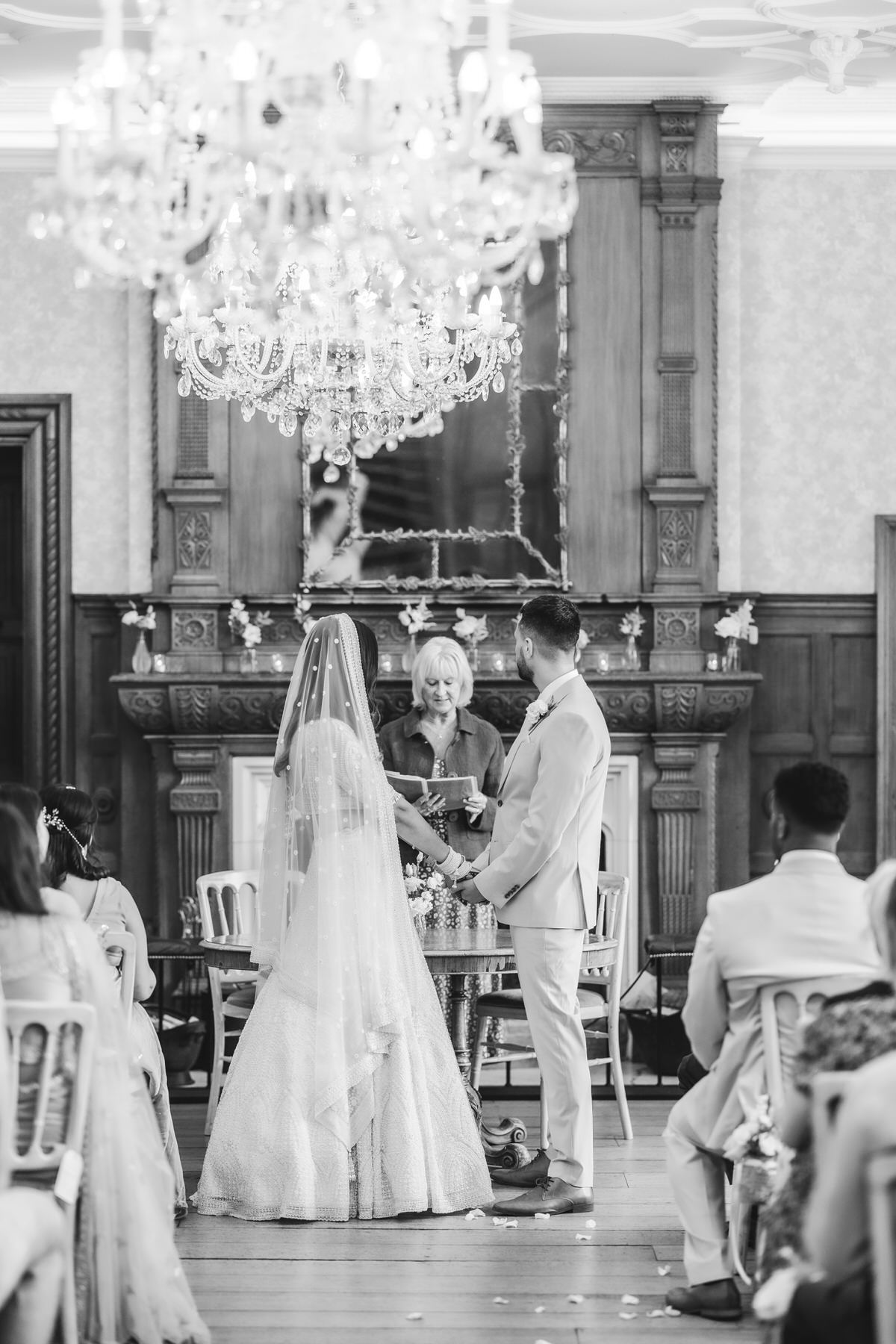 A bride and groom stand facing each other at an indoor wedding ceremony, holding hands beneath a large ornate chandelier, with guests seated around them and an officiant standing behind. The image is in black and white.