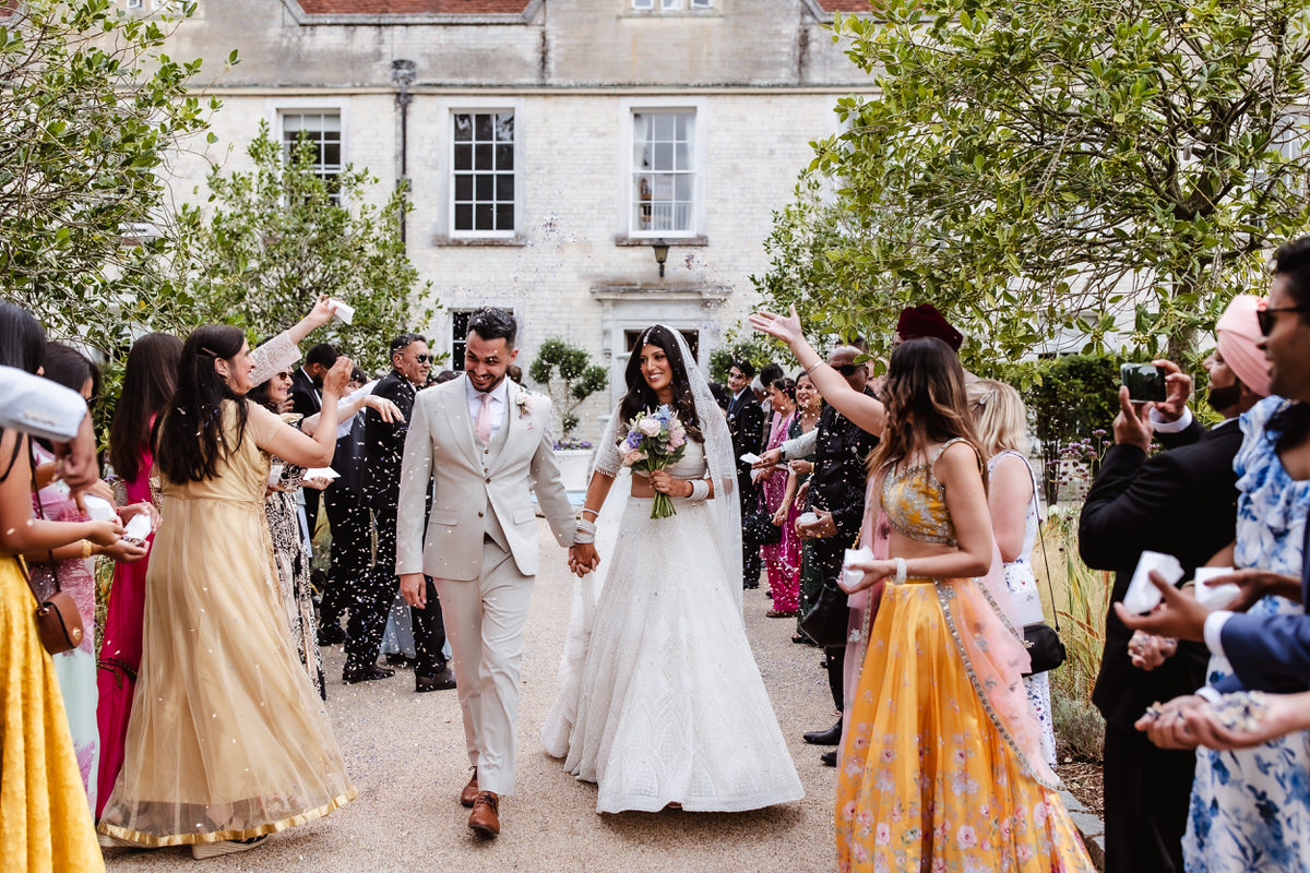 A newlywed couple walks hand in hand, smiling, as guests on both sides throw confetti at them outdoors. The bride wears a white dress and holds a bouquet; the groom wears a light suit. Trees and a historic building are in the background.
