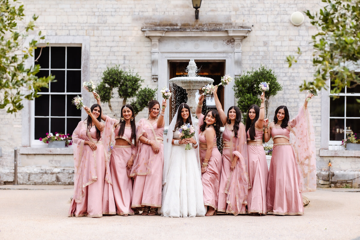 A bride in a white gown stands with her bridesmaids, who wear matching pink outfits, all holding bouquets and raising one arm in celebration in front of a white building with greenery and a fountain.