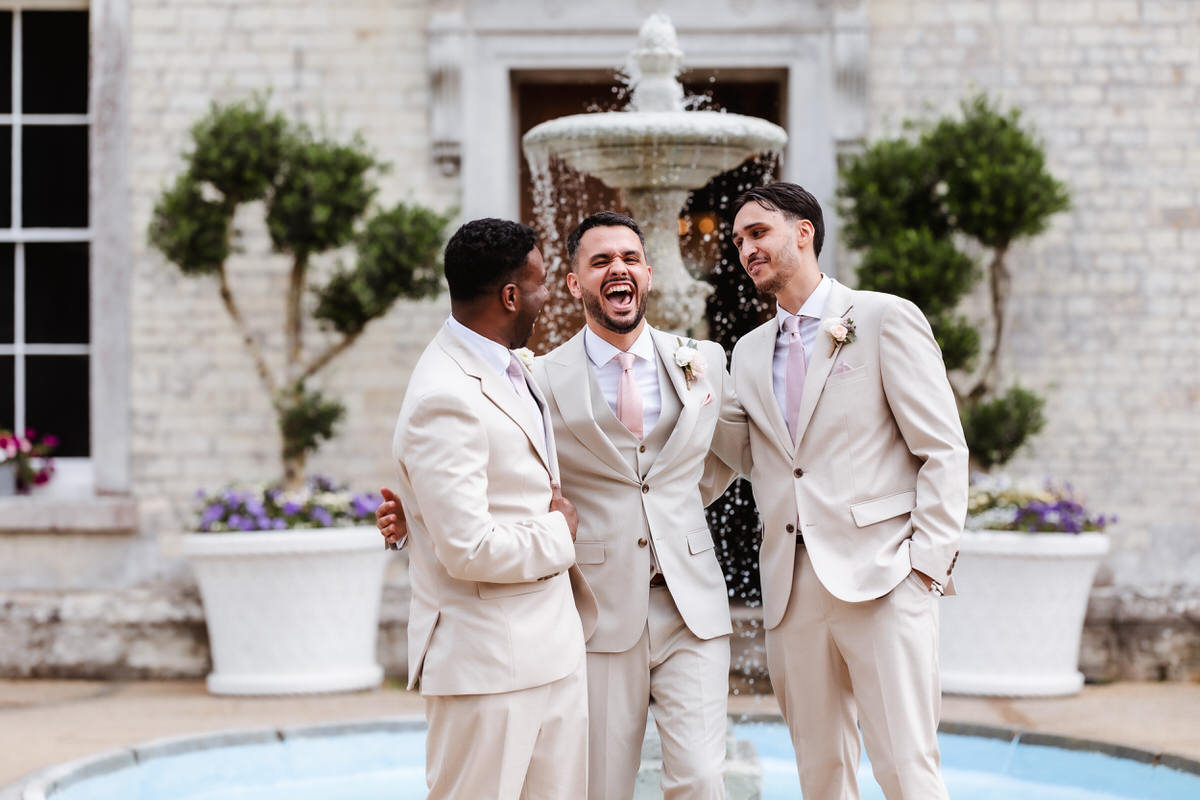 Three men in light beige suits stand laughing together in front of a fountain outside a building, enjoying a joyful moment during what appears to be a formal event or wedding.