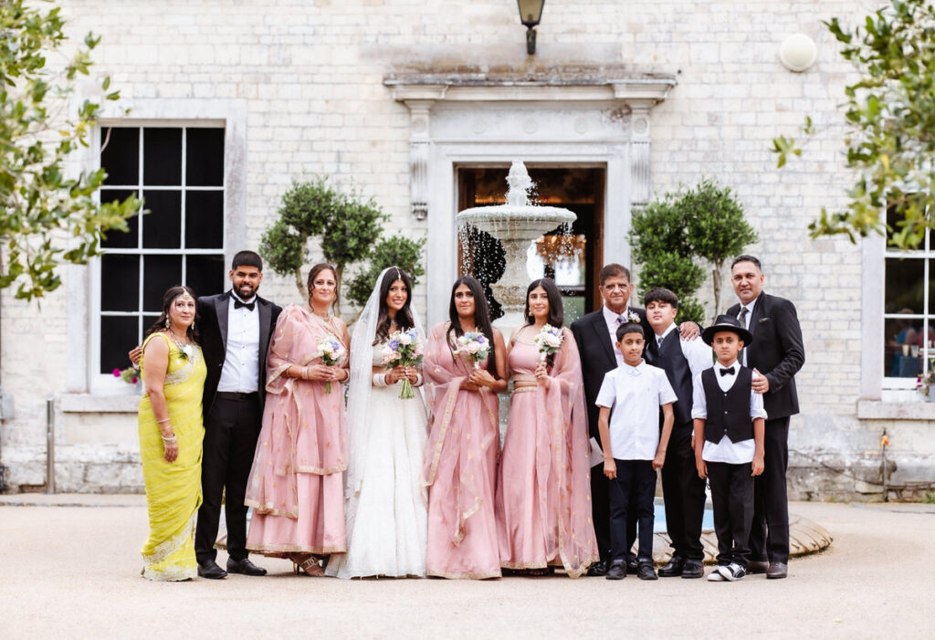 A wedding party poses outdoors in front of a stone building and a fountain. The group includes women in pink and gold dresses, men and boys in suits, and one woman in a yellow sari, all smiling at the camera.