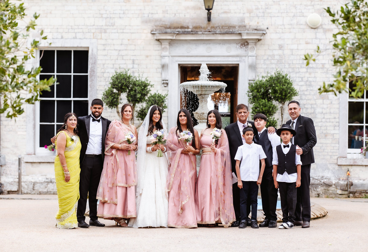 A wedding party poses outdoors in front of a stone building and a fountain. The group includes women in pink and gold dresses, men and boys in suits, and one woman in a yellow sari, all smiling at the camera.