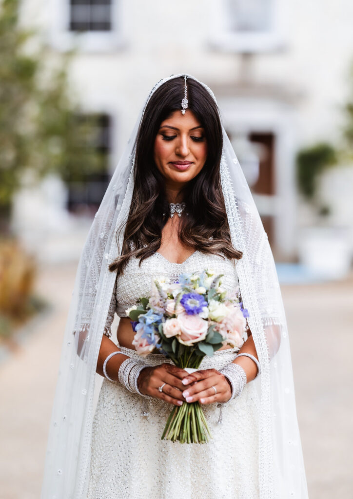A bride in a white, embellished outfit and veil holds a bouquet of pastel flowers, standing outdoors. She wears traditional jewelry and looks down, smiling softly. The background is blurred.