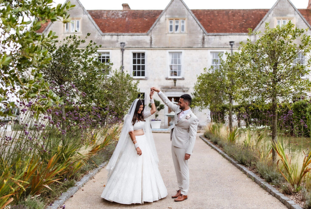 A bride in a white dress and a groom in a light suit dance joyfully on a garden path in front of a historic stone building, surrounded by greenery and flowering plants.