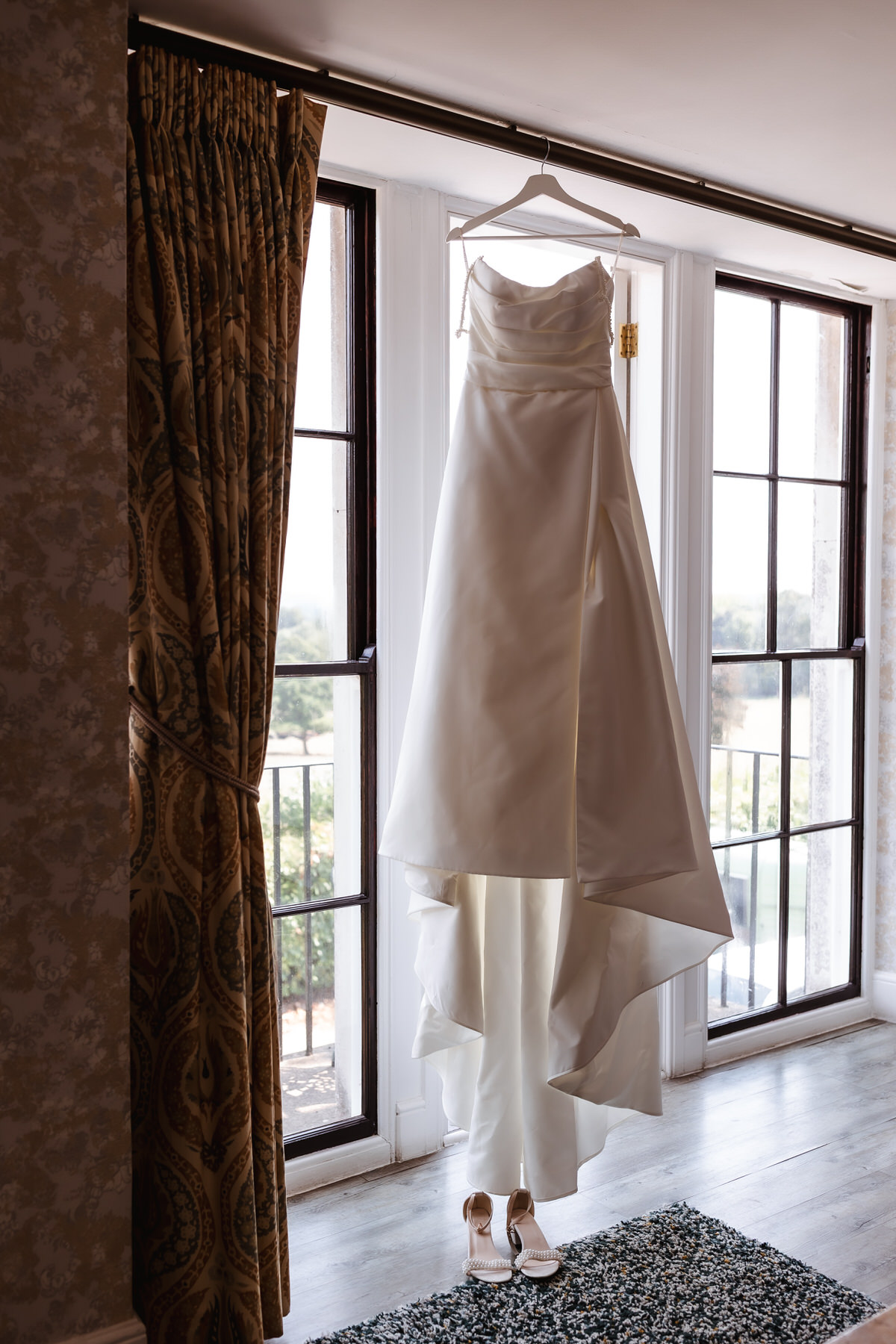 A white strapless wedding dress hangs on a wooden hanger in front of tall windows, with a pair of beige high-heeled shoes placed on the floor below. Sunlight softly illuminates the scene.