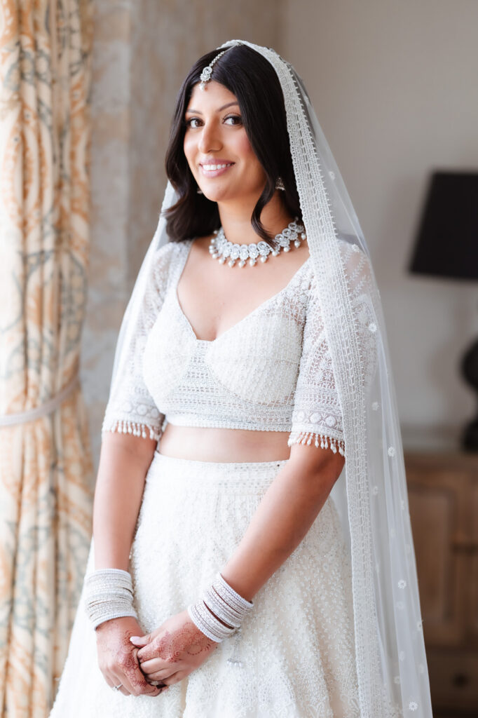 A smiling bride in a white embroidered lehenga and dupatta stands indoors by a window. She wears traditional jewelry, including a necklace, maang tikka, and bangles, with henna on her hands.