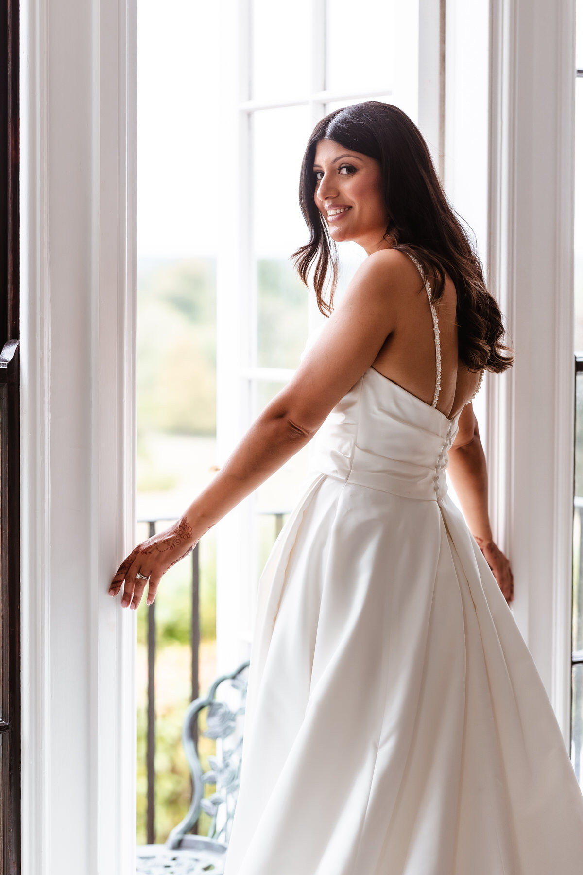A woman in a white, sleeveless wedding dress stands by tall windows, smiling and looking over her shoulder. Soft natural light illuminates her, highlighting her long dark hair and elegant gown.
