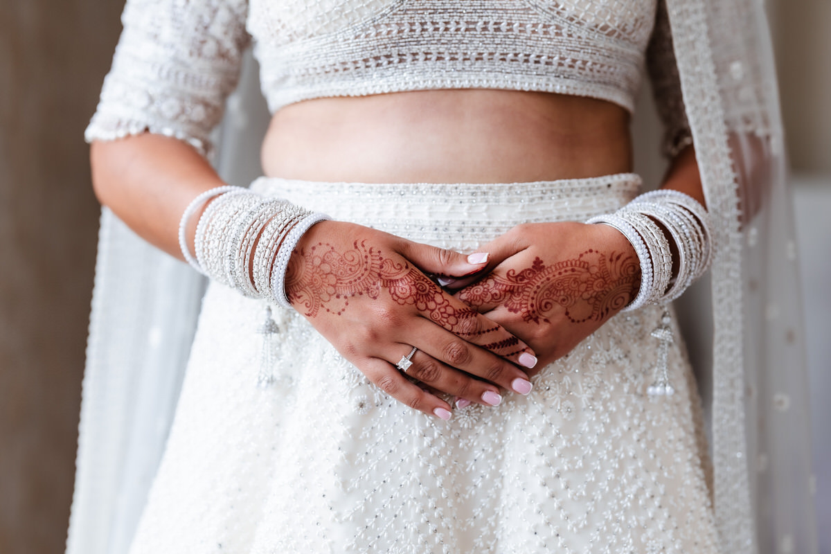 A woman in an embellished white outfit shows intricate henna designs on her hands, which are adorned with white bangles and a wedding ring. Her nails are painted light pink, and her midriff is visible.