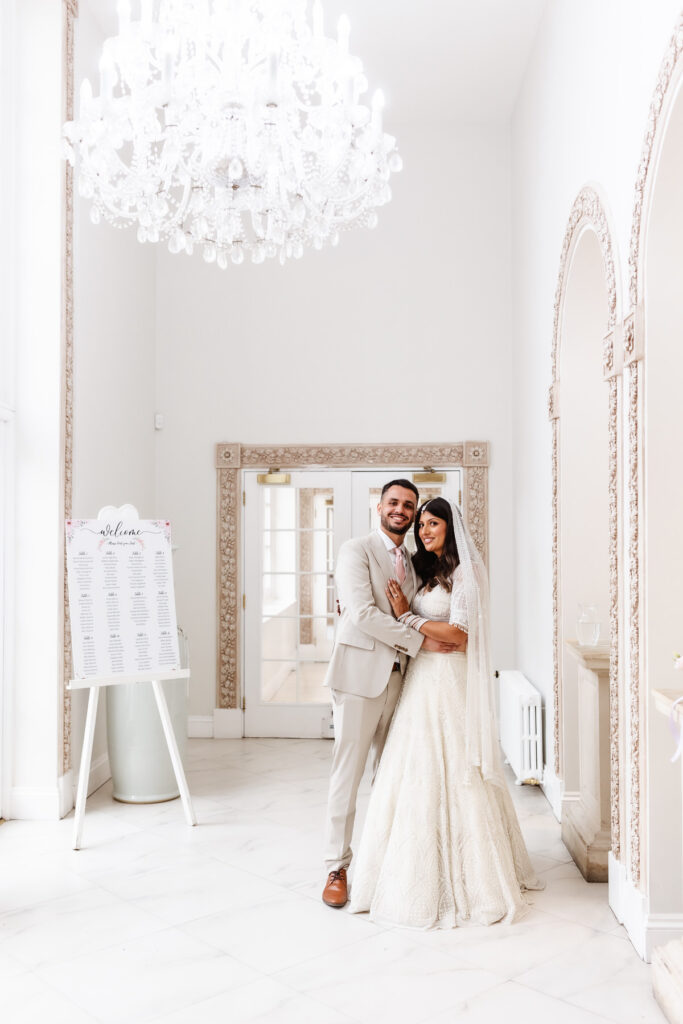 A bride and groom stand together, smiling and embracing in an elegant, white, chandelier-lit room with ornate trim and a wedding seating chart on display. The bride wears a lace gown and veil; the groom wears a light suit.