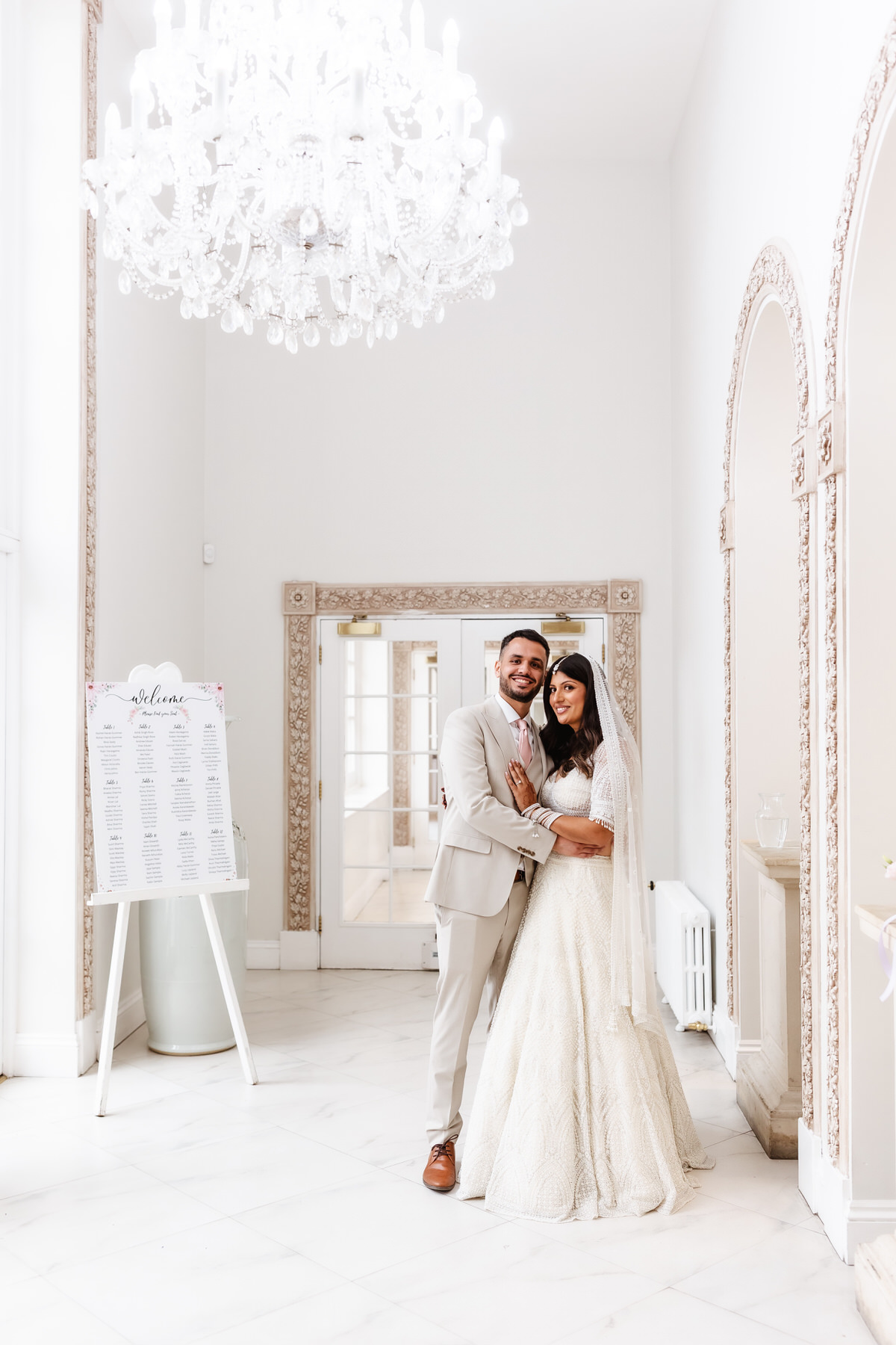 A bride and groom stand together, smiling and embracing in an elegant, white, chandelier-lit room with ornate trim and a wedding seating chart on display. The bride wears a lace gown and veil; the groom wears a light suit.
