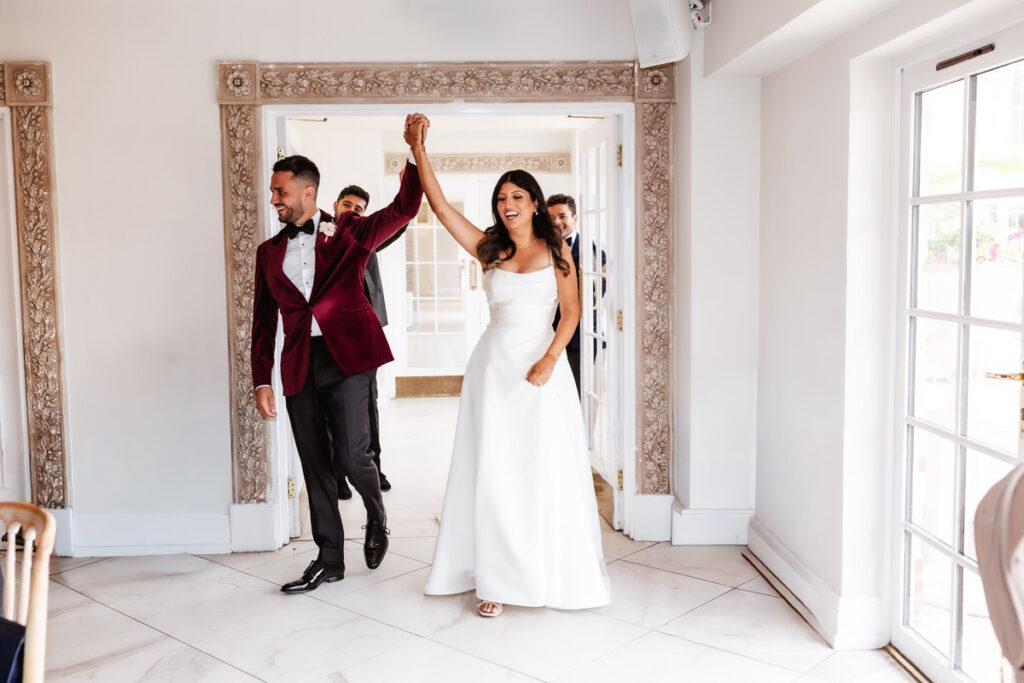 A bride in a white gown and a groom in a burgundy tuxedo jacket enter a bright room hand in hand, smiling as they celebrate their wedding.