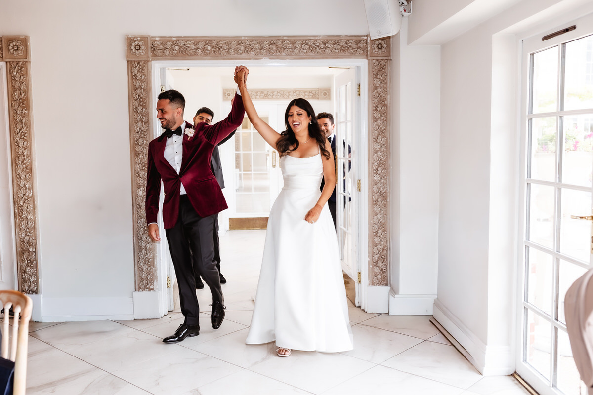A bride in a white gown and a groom in a burgundy tuxedo jacket enter a bright room hand in hand, smiling as they celebrate their wedding.