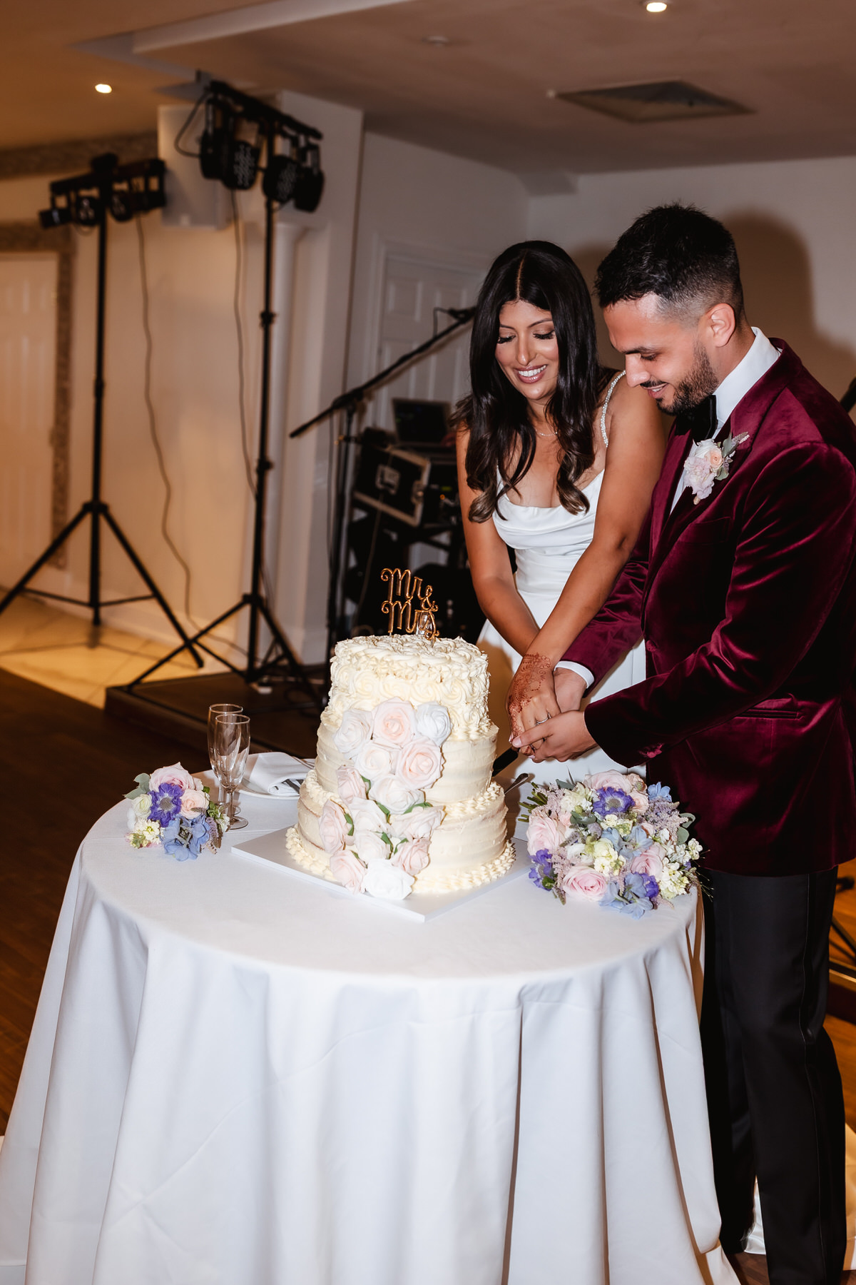 A bride and groom smile as they cut a decorated wedding cake together at a round table adorned with floral arrangements and champagne glasses.