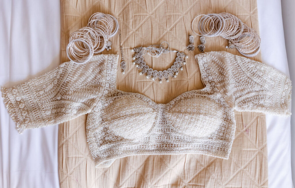 A cream-colored, intricately embroidered blouse is laid out with matching silver jewelry, including a necklace, earrings, and multiple bangles, arranged neatly on a beige textured cloth.