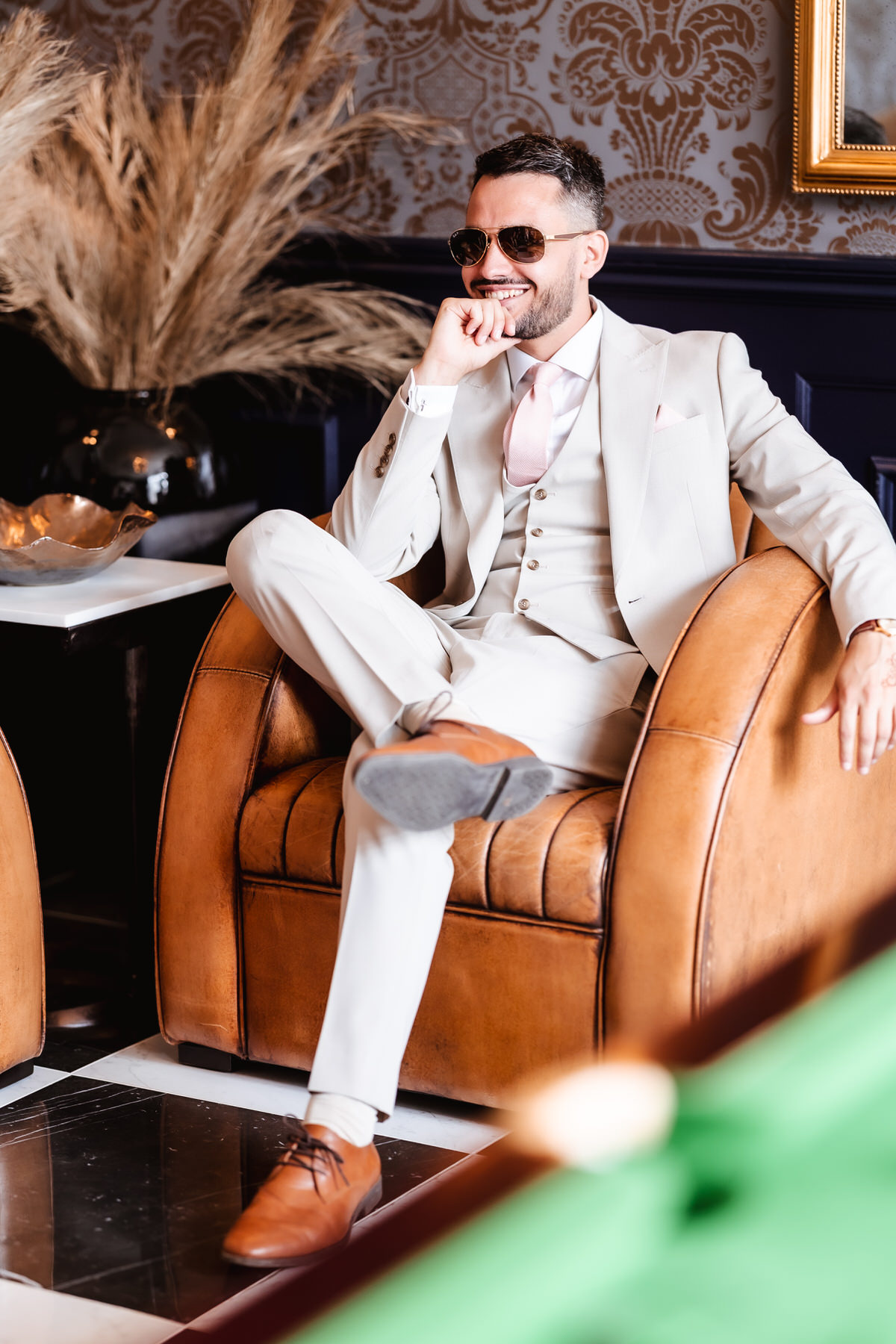A man in a light-colored suit and sunglasses sits confidently on a brown leather chair, smiling with one hand to his face. Elegant decor with pampas grass and patterned wallpaper is visible in the background.