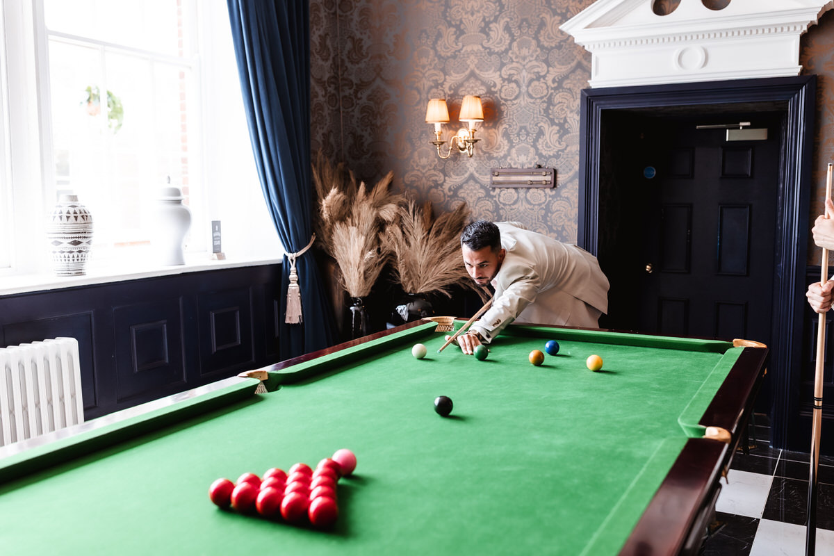A man in a light suit is aiming a shot at a green pool table in a stylish, well-lit room with patterned wallpaper, plants, and large windows. Another person is holding a cue at the edge of the table.