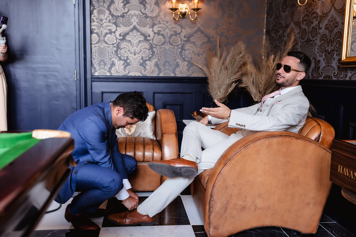 A man in a blue suit kneels to tie the shoe of a seated man in a light suit and sunglasses, who is relaxed with one arm outstretched, in a stylish room with leather chairs and decorative wallpaper.