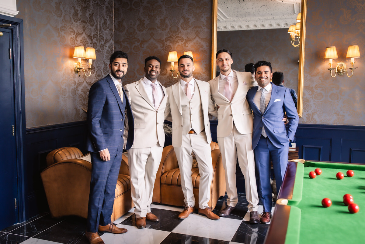 Five men dressed in suits, three in cream and two in blue, stand smiling together in an elegant room with patterned wallpaper, a large mirror, and a pool table with red balls in the foreground.