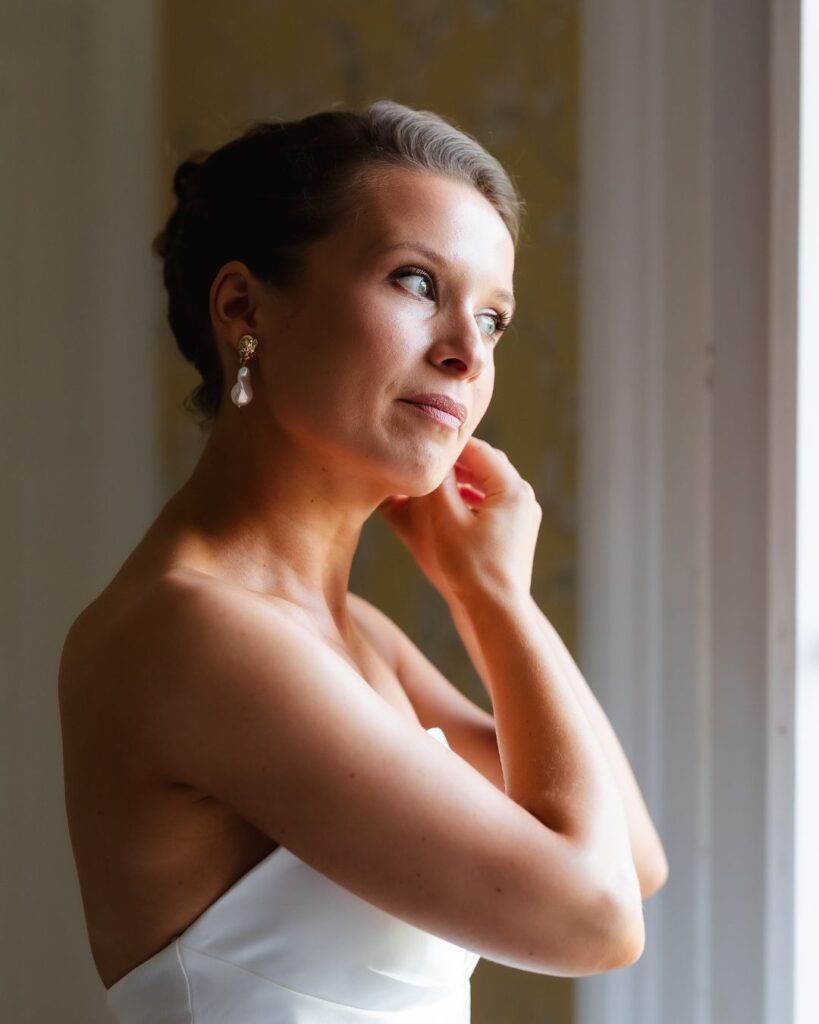 A woman in a strapless white dress stands by a window, touching her earring and gazing thoughtfully outside. Soft natural light highlights her face and elegant updo hairstyle.