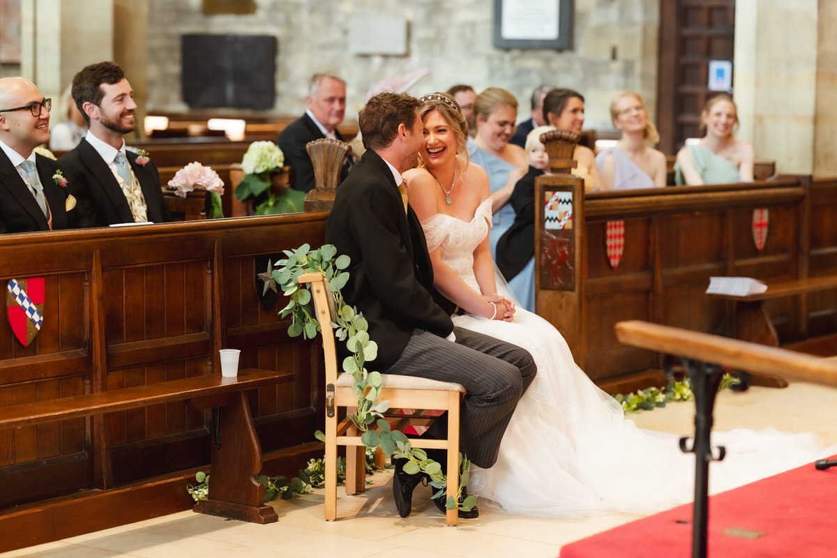 A bride and groom sit smiling and laughing together at the front of a church, surrounded by happy guests seated in wooden pews decorated with greenery. The atmosphere is joyful and celebratory.