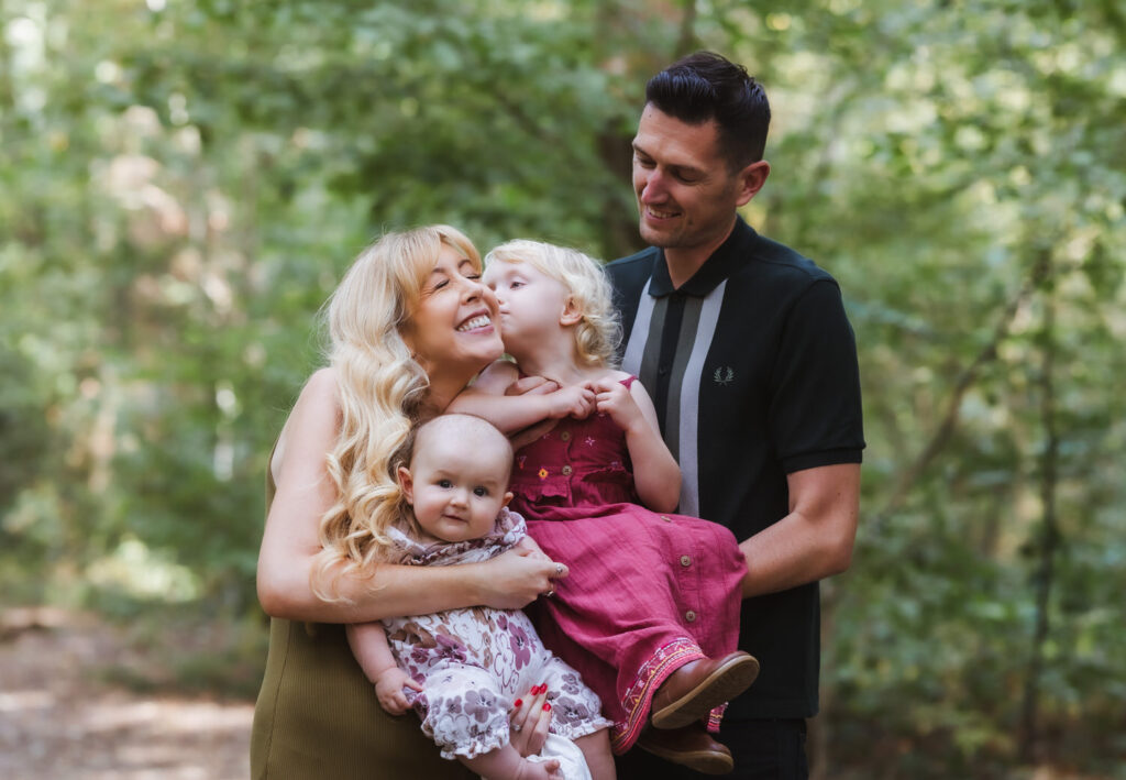 A smiling family of four stands outdoors. The woman holds a baby and a small child who kisses her cheek. The man stands beside them, smiling. They are surrounded by green trees.