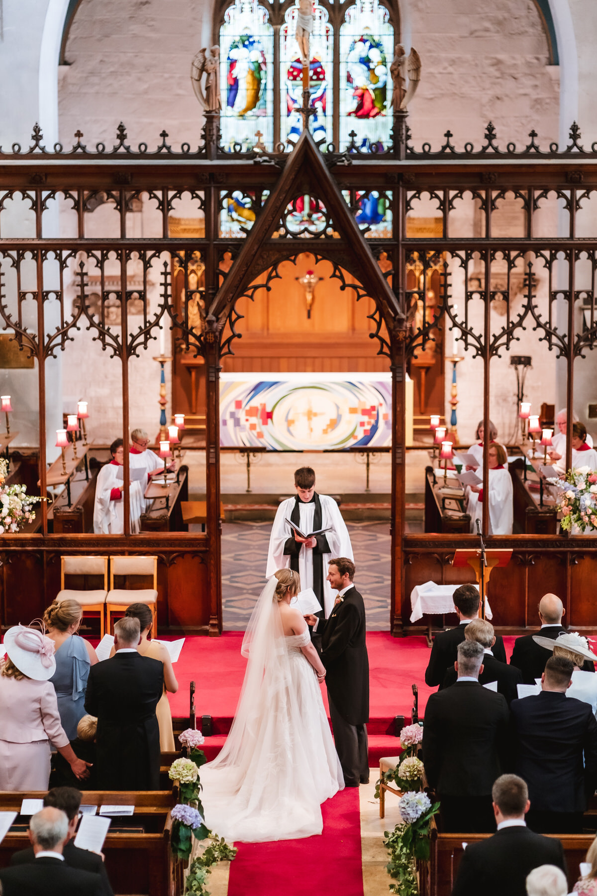 Bride and groom stand facing each other at the altar of a church during their wedding ceremony, surrounded by guests and a priest. The church is decorated with flowers and features stained glass windows.
