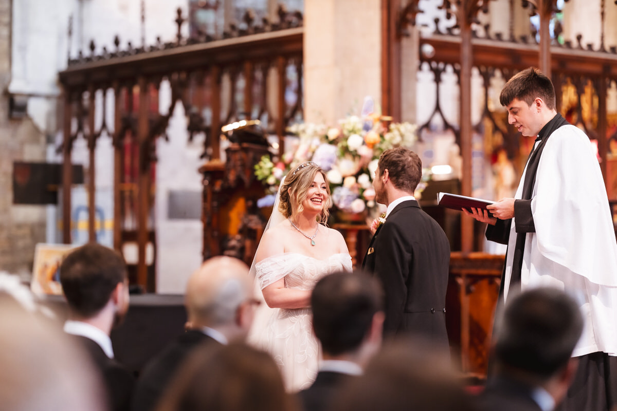 A bride and groom stand facing each other, smiling, during their wedding ceremony in a church. A priest in white robes stands beside them, reading from a book. Guests are seated in the foreground.