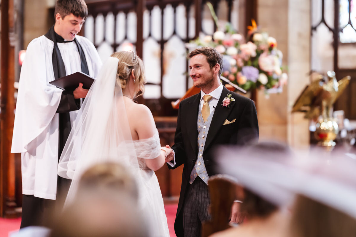 A bride and groom stand facing each other, holding hands and smiling during their wedding ceremony in a church, with an officiant in robes standing behind them and flowers in the background.