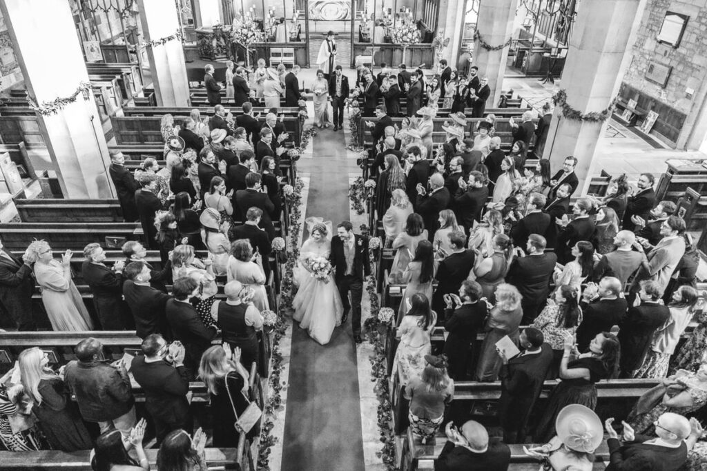 A bride and groom walk down the aisle of a crowded church, surrounded by guests who are standing, smiling, and taking photos. The image is taken from above, capturing the festive atmosphere of a wedding ceremony.