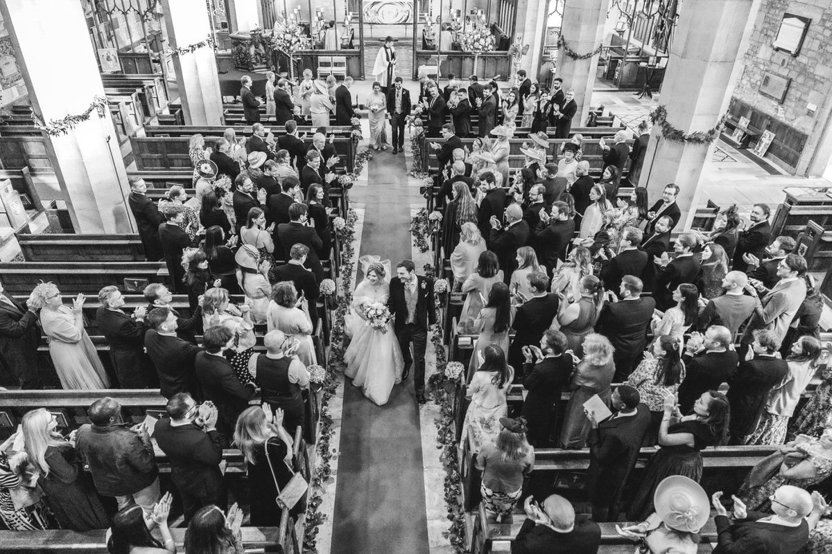 A bride and groom walk down the aisle of a crowded church, surrounded by guests who are standing, smiling, and taking photos. The image is taken from above, capturing the festive atmosphere of a wedding ceremony.