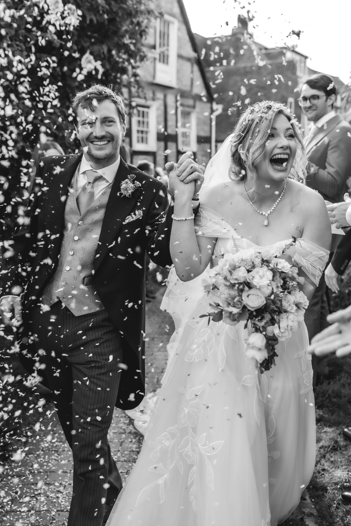 A joyful bride and groom walk hand in hand outdoors, surrounded by guests tossing confetti. The bride holds a bouquet and both are smiling widely, celebrating their wedding day. The image is in black and white.