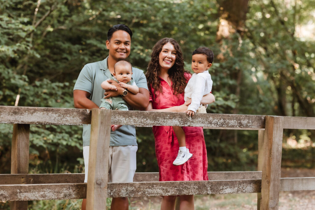 A smiling family of four stands on a wooden bridge in a lush green forest. The father holds a baby, while the mother, in a red dress, holds a toddler wearing white. Everyone looks happy and relaxed outdoors.