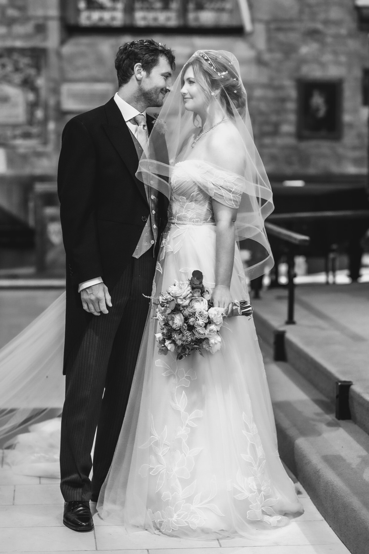 A bride and groom stand together in a church, smiling at each other. The bride wears a veil and holds a bouquet of flowers; the groom is in a suit. The image is black and white.