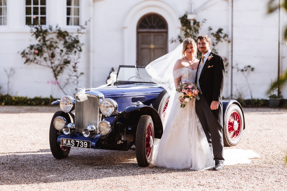 A bride and groom stand smiling beside a vintage blue convertible car with red wheels, parked in front of a white building. The bride holds a bouquet and wears a veil; the groom is in a black suit.